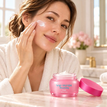 Woman applying white cream to her cheek beside an open pink NAD+ Peptide Boosting Cream jar and matching lid.